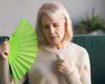 Overheated elderly woman sitting on couch waving green fan to cool herself, sixty years female feels unwell hot, age hormonal changes, apartments without air conditioner, summertime discomfort concept