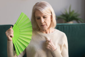 Overheated elderly woman sitting on couch waving green fan to cool herself, sixty years female feels unwell hot, age hormonal changes, apartments without air conditioner, summertime discomfort concept