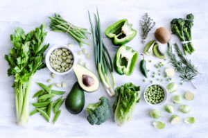 A variety of green and leafy vegetables and fruits, of which some are cut in half. Included in the variety of vegetables and fruits is an avocado, green bell peppers, kiwis, peas, beans and lettuce. They are placed in a rustic white background.