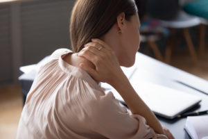 Back view of young woman touch neck suffer from sudden muscular spasm at workplace, unwell millennial female worker massage upper back having backache or pinched nerves sitting in incorrect posture