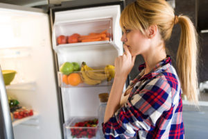 Shot of pretty young woman hesitant to eat in front of the fridge in the kitchen.