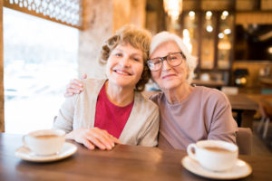 Cheerful positive attractive senior lady friends sitting at table with cups and embracing while looking at camera in coffee shop
