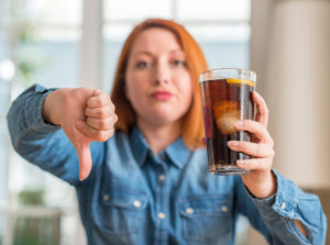 Redhead woman holding soda refreshment with angry face, negative sign showing dislike with thumbs down, rejection concept