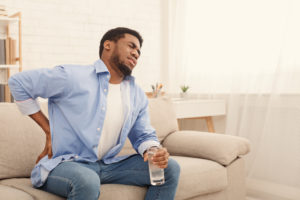 Young african-american man with back pain, pressing on hip with painful expression, sitting on sofa at home with glass of water, copy space
