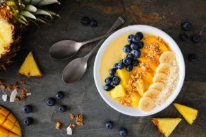 Healthy pineapple, mango smoothie bowl with coconut, bananas, blueberries and granola. Top view table scene on a dark background.