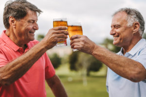 Two senior men toasting beer glasses outdoors. Smiling mature male friends cheering beers while standing outside.