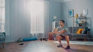 Muscular Athletic Fit Man in T-shirt and Shorts is Doing Squat Exercises at Home in His Spacious and Bright Living Room with Minimalistic Interior.
