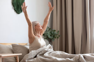 Happy older woman sitting in comfortable bed, stretching hands after awakening in bedroom, happy satisfied older female doing easy exercise in the morning, greeting new day, sunrise