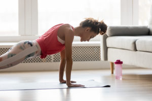 Young attractive sportive woman wearing activewear doing push ups or press ups exercise position on sport mat at home, practicing yoga plank pose, working out indoors, healthy active lifestyle concept