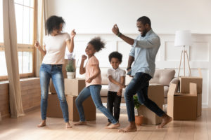 Happy african american parents and cute children dancing among boxes celebrating moving day relocation renovation, active carefree funny mixed race family mom dad having fun with kids in new house