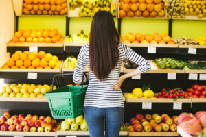 Back view of a young woman in casual clothes with a basket for food in hand on a background of fruits and vegetables in a supermarket.