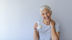 Cheerful mature woman having fun while drinking milk. Senior woman drinking from a clear glass full of milk. Woman in her golden age. Smiling, beautiful senior lady drinking a glass of milk