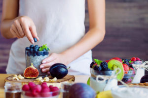 Female hands are preparing yogurt with chia and blueberries for good digestion, functioning of gastrointestinal tract. Summer berries, nuts, fruits, dairy products on table. Healthy food concept.