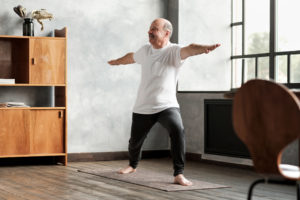 Senior hispanic man standing in warrior two yoga pose practicing in living room alone