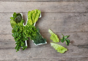 Vitamin K in food concept. Plate in the shape of the letter K with different fresh leafy green vegetables, lettuce, herbs on wooden background. Flat lay or top view.