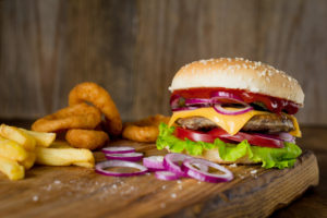 Cheeseburger, french fries and onion rings on wooden chopping board over wooden backdrop. Horizontal, closeup view