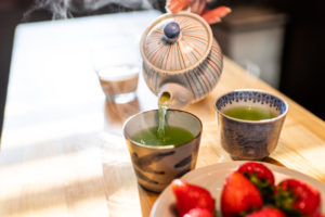 Woman hand holding teapot pouring hot traditional Japanese sencha green tea into cup with dessert strawberries on plate on at wooden kitchen table with steam