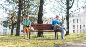 Woman and man with face mask in social distancing flirting sitting on a park bench