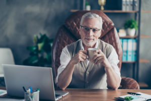 Cheerful careless excited joyful old businessman with bristle is pondering about holidays sitting on the armchair in front of laptop