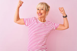 Middle age woman wearing striped t-shirt standing over isolated pink background showing arms muscles smiling proud. Fitness concept.