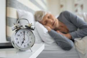 Closeup of alarm clock with senior woman in deep sleep at home. Old woman sleeping in bed next to alarm clock in morning. Elderly woman sleeping in bedroom peacefully.