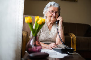 Old woman with gray white hair and glasses sitting in her armchair in her home and talking on the phone. Grandmother is happy to talk to her children and grandchildren.