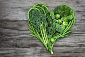 Green vegetables in a heart shape on an old wood background. Vegetables include various lettuce, kale, green beans, asparagus, brussel sprouts, and broccoli.