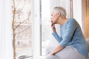 Mindful mature woman sitting by the window in her home and smoking cigarette, Unhealthy habits concept