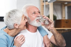 Happy beautiful senior man, white beard and grey hair, holding transparent glass in his hand and drink water. Smiling wife near by. Concept healthy life and diet for senior people. Closeup.