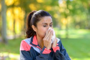 Woman blowing her nose on a tissue outdoors in a leafy green park while out jogging conceptual of seasonal flu or allergies