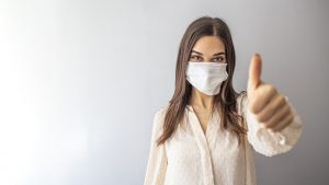 Beautiful caucasian young woman with disposable face mask. Protection versus viruses and infection. Studio portrait, concept with white background. Woman showing thumb up.