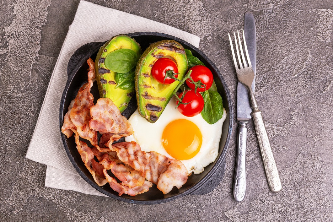 Grilled bacon and avocado, fried eggs with spinach and cherry tomatoes in cast-iron pan. Gray concrete background. Top view. Ketogenic diet. Low carb high fat breakfast. Healthy food concept