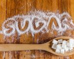 The word sugar written into a pile of white granulated sugar with spoon of sugar cubes over wooden background