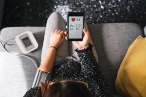 Young woman measures blood pressure sitting on sofa at home with smartphone connected to device - Concept of health, well-being and love for oneself - Millennial in a moment of private life