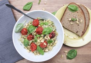 Orzo pasta with pesto, cherry tomatoes, pine nuts and basil on top of a wooden table and with a loaf of bread on the side.