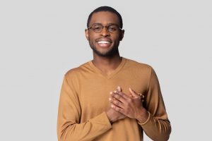 Head shot portrait close up smiling African American man in glasses keeping hands on chest, looking at camera, grateful young male feeling gratitude, appreciation, isolated on grey background