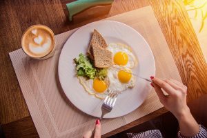 Top view of female hands holding fork and knife while having tasty breakfast at modern european cafe. Young woman eating fried eggs with toasts and morning coffee at restaurant