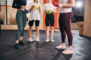Cropped photo of four slim young women in sporty gear holding resistance bands at the gym