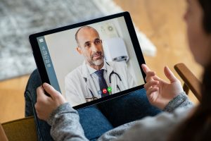 Back view of young woman making video call with her doctor while staying at home. Close up of patient sitting on armchair video conferencing with general practitioner on digital tablet. Sick girl in online consultation with a mature physician.