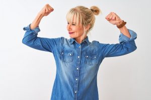 Middle age woman wearing casual denim shirt standing over isolated white background showing arms muscles smiling proud. Fitness concept.