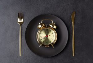 Golden alarm clock on a black plate with a golden knife and fork. Intermittent fasting concept. Horizontal orientation, top view.