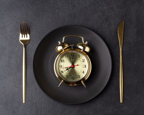 Golden alarm clock on a black plate with a golden knife and fork. Intermittent fasting concept. Horizontal orientation, top view.