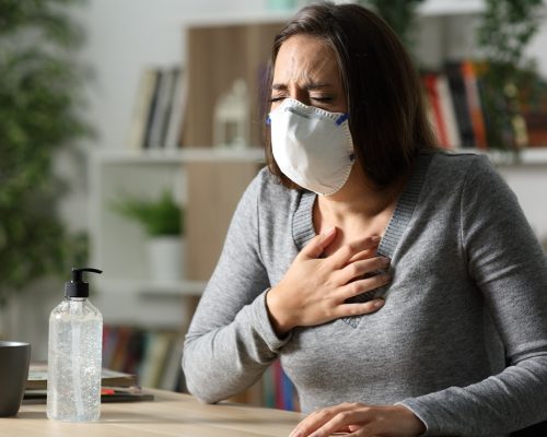 Woman with protective mask suffocating due coronavirus holding chest sitting on a desk at nigh at home
