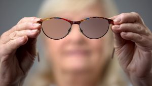 Old woman holding trendy glasses, concept of vision problems, blurred background