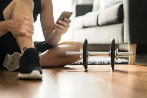 Man using smartphone during workout at home. Online personal trainer or on mobile phone. Internet fitness class or video course. Taking a break. Lazy guy with cellphone while training with dumbbell.