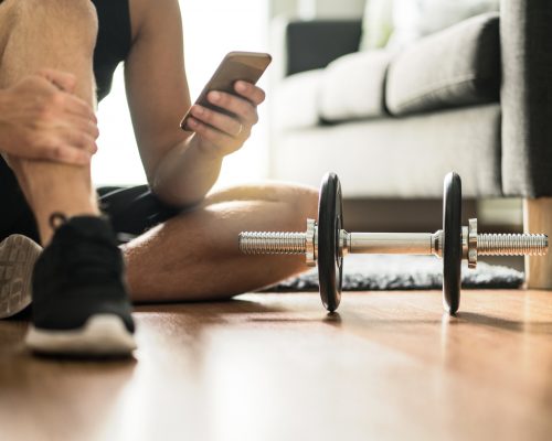 Man using smartphone during workout at home. Online personal trainer or on mobile phone. Internet fitness class or video course. Taking a break. Lazy guy with cellphone while training with dumbbell.