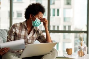 African American woman with face mask having a headache while going through paperwork and using laptop while working at home.