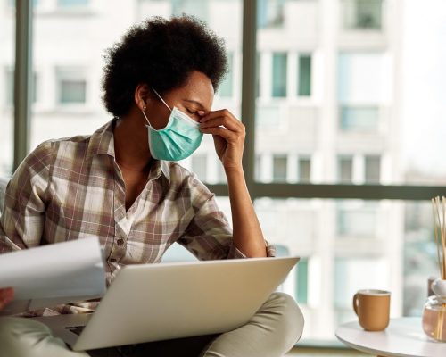 African American woman with face mask having a headache while going through paperwork and using laptop while working at home.