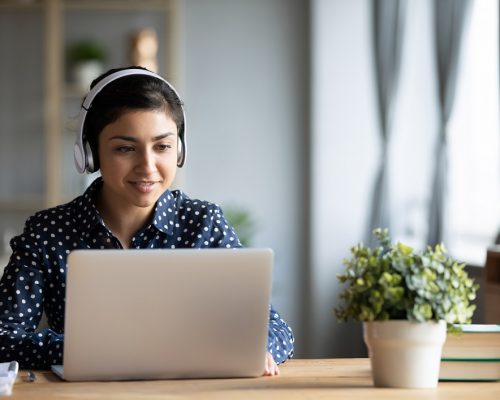 Millennial Indian girl in wireless headphones sit at desk at home working on modern laptop, young ethnic woman in earphones browsing Internet shopping online or studying on computer in living room