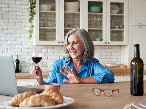 Happy 60s mature woman drinking wine video calling friend on laptop at home. Smiling old middle aged lady holding glass waving hand talking by online social distance chat meeting sit at kitchen table.